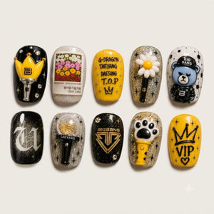 A set of ten decorative press-on nails arranged in two rows on a light beige background. The nails feature various designs including Bigbang signatures